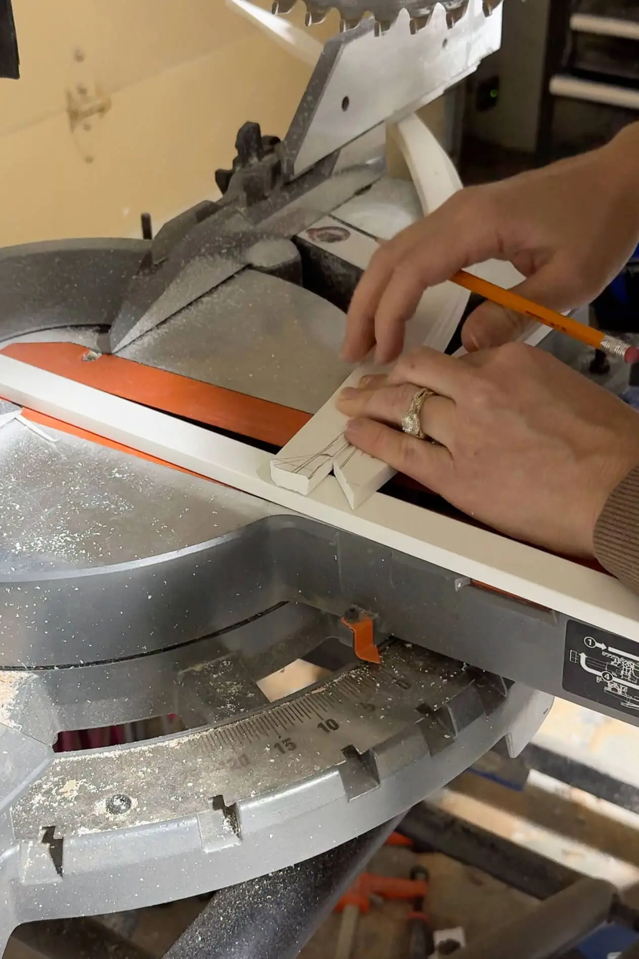 Close-up of hands measuring and marking a piece of white pvc trim on a miter saw, with sawdust scattered on the base and the blade angled for a precise cut.